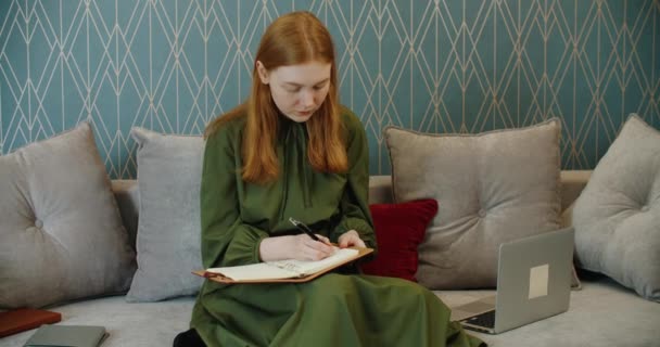 Vacker kvinna skriver anteckningar i en anteckningsbok sitter på soffan hemma. — Stockvideo
