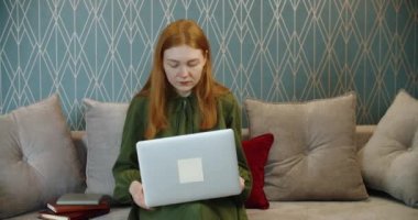 Evdeki kanepede çalışan genç bir kadın dizüstü bilgisayarda yazı yazıyor ve internetten alışveriş yapıyor..