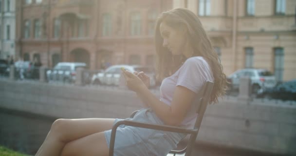 Gelukkige vrouw zit in het park en leest het nieuws op een mobiele telefoon. Park en huizen op de achtergrond. Portret van een gelukkige vrouw — Stockvideo