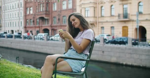 Mulher feliz senta-se no parque e lê as notícias em um telefone celular. Parque e casas no fundo. Retrato de uma mulher feliz — Vídeo de Stock