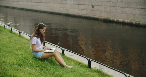 Mulher morena feliz trabalhando em laptop ao ar livre sentado na grama no parque. Belo lugar para trabalhar. Freelance. — Vídeo de Stock