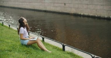 Dışarıda dizüstü bilgisayarda çalışan mutlu esmer kadın parkta çimlerin üzerinde oturuyor. Çalışmak için güzel bir yer. Bağımsız.