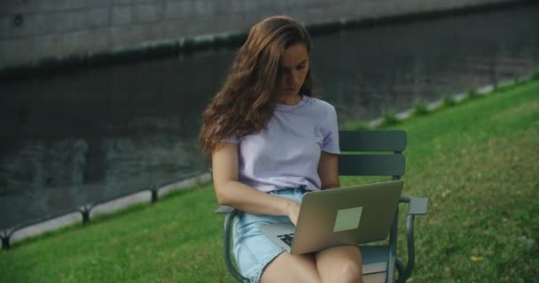 Щаслива брюнетка працює на ноутбуці на відкритому повітрі, сидячи на траві в парку. Гарне місце для роботи. Фріланс . — стокове відео