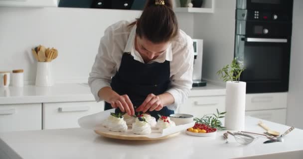 Приготовление и украшение десертов свежими ягодами. Молодая женщина-кондитер на кухне украшает десерт ягодами. Счастливая женщина в фартуке на кухне делает прекрасный десерт на день рождения — стоковое видео