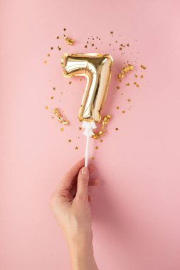 Pembe arka plandaki çubuk altın konfetisinin üzerinde 7 numaralı şişirilebilir altını tut. Tatil, doğum günü, yıldönümü kavramı.