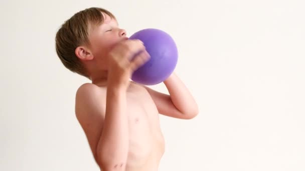 Un niño intenta inflar un globo púrpura sobre un fondo blanco. — Vídeos de Stock