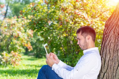 Ağacın altında tablet bilgisayar kullanan genç adam.