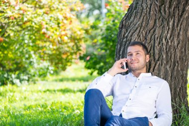 Genç adam ağacın altında akıllı telefondan konuşuyor.