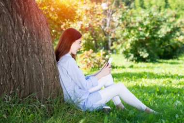 Genç kızıl saçlı kadın ağacın altında akıllı telefon kullanıyor.