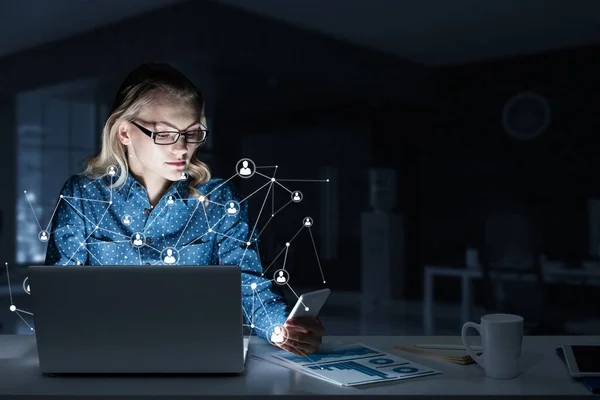 Loira atraente usando óculos no escritório escuro usando laptop. Meios mistos — Fotografia de Stock