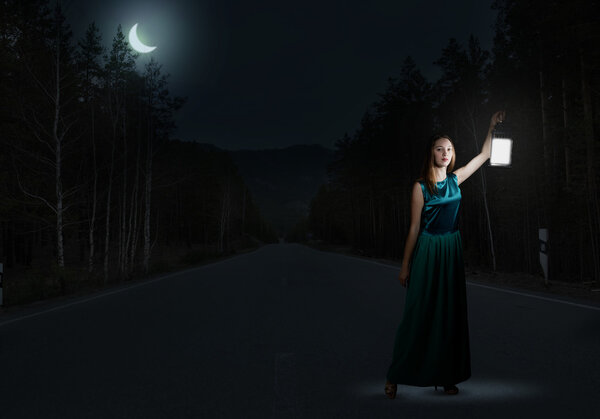 Girl lost in night. Young attractive woman in green dress with lantern walking in darkness