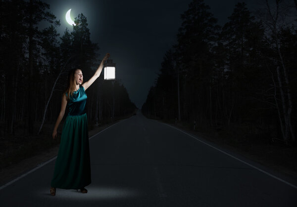 Girl lost in night. Young attractive woman in green dress with lantern walking in darkness
