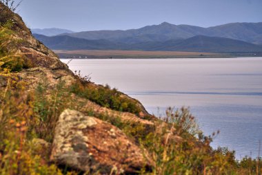 High-angle drone shot of a desolate coastal landscape featuring a narrow sandy beach, winding dirt tracks, and small campsites. The deep blue water surrounds a rocky peninsula and islands, with a dramatic mountain range under an overcast, moody sky i