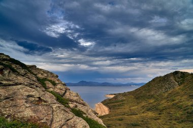 High-angle drone shot of a desolate coastal landscape featuring a narrow sandy beach, winding dirt tracks, and small campsites. The deep blue water surrounds a rocky peninsula and islands, with a dramatic mountain range under an overcast, moody sky i