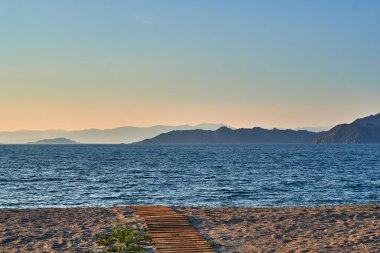 A stunning landscape shot of the sun setting behind the rugged mountain ridges of the Bukhtarma Reservoir. The image features a bright orange sky reflecting its warm glow on the water surface and creating dark mountain silhouettes, capturing the peac