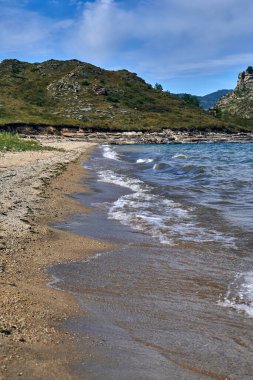 Issız bir sahil manzarasının yüksek açılı drone görüntüsü. Dar kumlu bir sahil, virajlı toprak yollar ve küçük kamp alanları. Derin mavi su, bulutlu ve kasvetli bir gökyüzünün altında dramatik bir sıradağla kayalık bir yarımadayı ve adaları kaplar.