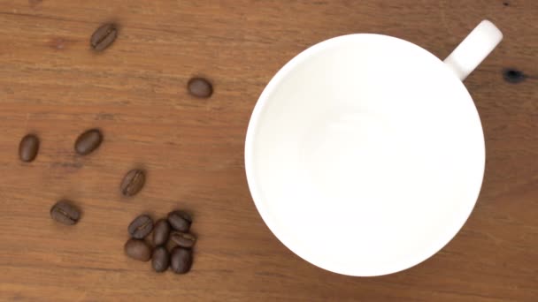 金属製のスプーンから木製のテーブルの上に焙煎されたコーヒー豆と白いセラミックコーヒーカップに落ちる初期のコーヒー粉のトップビュー — ストック動画