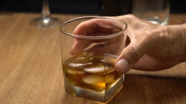 老人はウイスキーグラスをアイスキューブで持ち上げ 木製のテーブルの上に置きます — ストック動画