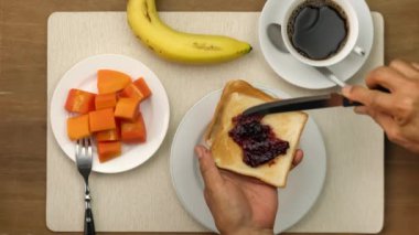 Kahvaltıda bir tostun üzerine bıçak sürüp reçel sürerken bir fincan sıcak sade kahve, olgun muz ve olgun papaya parçalarıyla yaşlı adamın üst görüntüsü..