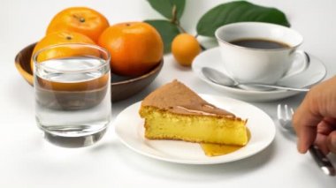 Bir bardak su, bir fincan sade kahve, ahşap kasede portakallar ve beyaz arka planda olgun bir Marian eriği ile ev yapımı ballı turuncu pandispanyadan bir ısırık alarak..