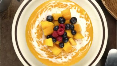 Olgun mango, ahududu, yabanmersini, üzüm ve krema parçalarıyla süslenmiş ev yapımı mango peynirli kek kahverengi seramik tabakta döner ve ahşap masaya yakınlaşır..