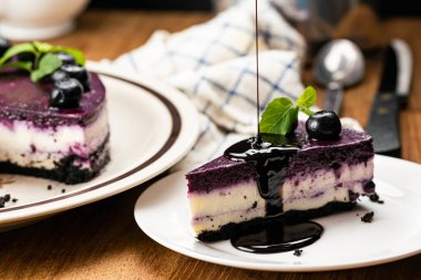 Lezzetli ev yapımı yabanmersinli cheesecake parçasına çikolata dökerek yaban mersinli ve naneli beyaz seramik tabakla süslenmiş. Kahverengi tabakla, beyaz kahve fincanıyla, metal kaşıkla ve bıçakla ahşap masada..