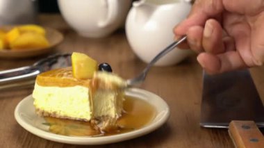 Metal kaşık kullanarak lezzetli ev yapımı mango cheesecake yemek ahşap masadaki seramik tabakta balla birlikte kürek, kahve makinesi ve ahşap masada süt demliği..
