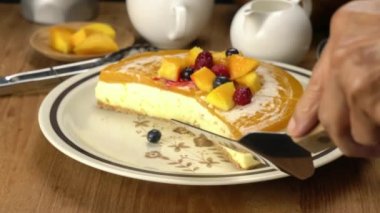 Sunucu küreği kullanan usta bir parça lezzetli ev yapımı mango peynirli kek al olgun mango parçaları, korunmuş ahududu ve yabanmersini ile süslenmiş ahududu ve ahşap tabakta kahverengi seramik tabakta.
