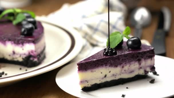 将巧克力倒在一块自制蓝莓芝士蛋糕上 用保存好的蓝莓和薄荷装饰在白色陶瓷盘子里 整个盘子都是褐色的 刀叉和勺子放在木制桌子上 — 图库视频影像