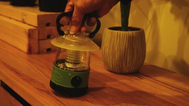Modern batarya portatif bir LED kamp feneri getir, ahşap bir masaya koy ve çalıştır..