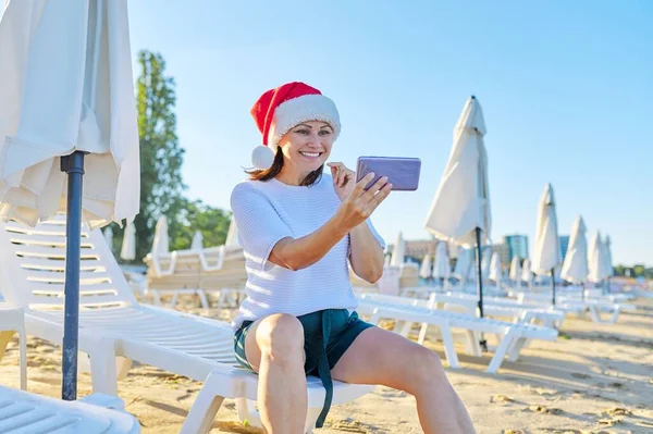 Πρωτοχρονιάτικες και Χριστουγεννιάτικες διακοπές, ευτυχισμένη γυναίκα με καπέλο Σάντα στην παραλία κάνοντας βιντεοκλήση — Φωτογραφία Αρχείου