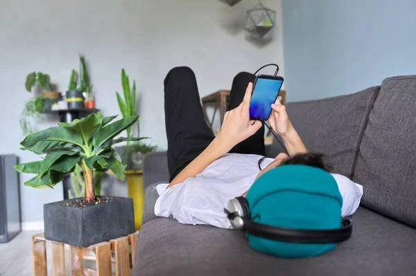 Smartphone pantalla azul en las manos del hipster adolescente acostado en el sofá con sombrero con auriculares —  Fotos de Stock