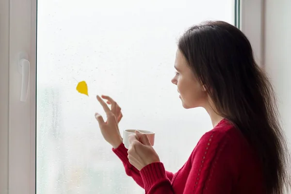 Портрет женщины, смотрящей в окно с каплями дождя, осенний желтый лист — стоковое фото