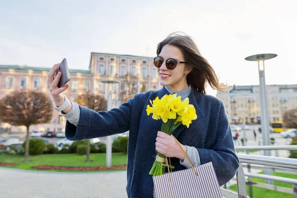 Piękna szczęśliwa młoda kobieta w płaszczu z telefonu z bukietem kwiatów na ulicy miasta — Zdjęcie stockowe