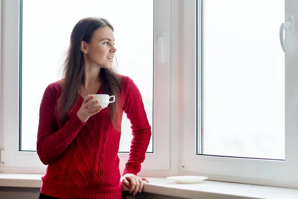 Ung, smilende kvinne som nyter kaffe, kvinne i varm, strikket genser ved vinduet – stockfoto