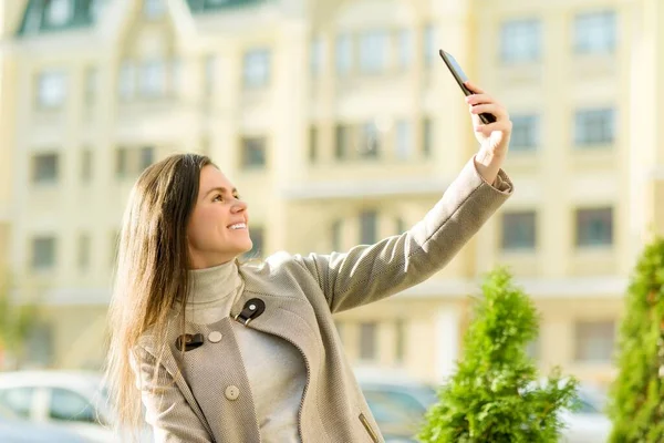 Piękna szczęśliwa młoda kobieta w płaszczu z telefonem na ulicy miasta — Zdjęcie stockowe