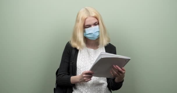 Menina adolescente estudante em máscara protetora médica em seu rosto, com mochila notebook, em fundo verde claro — Vídeo de Stock