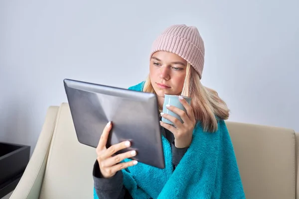 Έφηβος φοιτητής με ψηφιακή δισκίο φλιτζάνι τσάι στο καπέλο κάτω από ζεστή κουβέρτα κάθεται στο σπίτι — Φωτογραφία Αρχείου