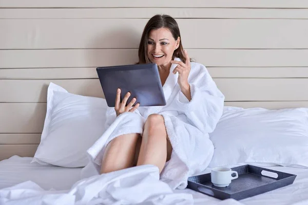 Зріла жінка в білому халаті з чашкою кави дивиться на веб-камеру цифрового планшета — стокове фото