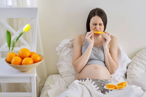 Jovem mulher grávida bonita comer laranjas sentado na cama em casa — Fotografia de Stock