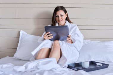Beyaz bornozlu, elinde bir fincan kahveyle yetişkin bir kadın dijital tabletin web kamerasına bakıyor.