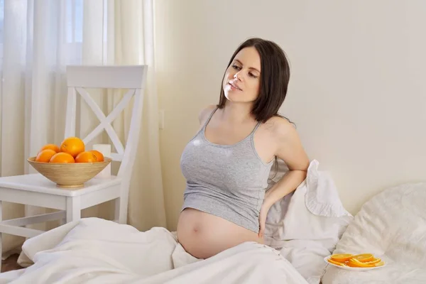 Mladá krásná těhotná žena s miskou pomerančů sedí v posteli doma — Stock fotografie