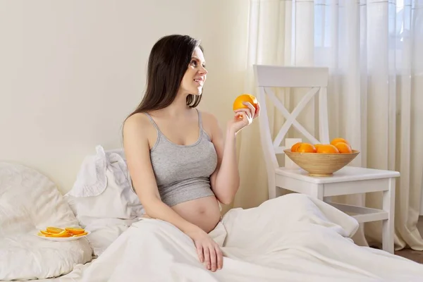 Jonge mooie zwangere vrouw eten sinaasappels zitten in bed thuis — Stockfoto