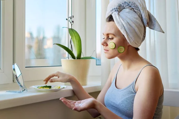 Jonge vrouw met badhanddoek op het hoofd van toepassing crème en komkommer plakjes op het gezicht — Stockfoto