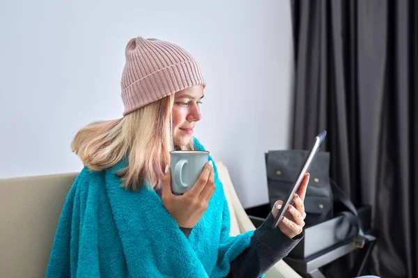 Adolescente estudiante con la tableta digital taza de té en el sombrero bajo una manta caliente sentado en casa —  Fotos de Stock