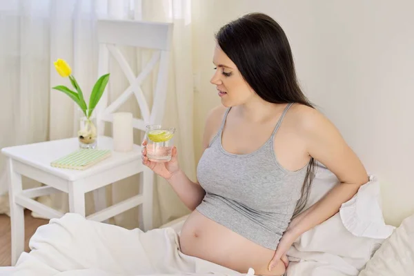 Krásná mladá těhotná žena pitná voda s citronem sedí doma v posteli — Stock fotografie