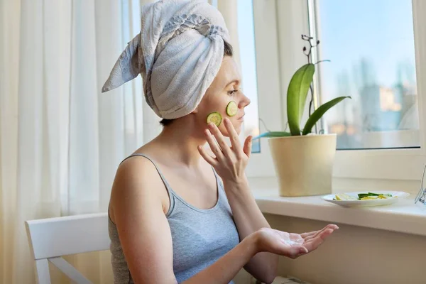 Jovem com toalha de banho na cabeça aplicando creme e fatias de pepino no rosto — Fotografia de Stock