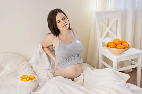 Mladá krásná těhotná žena s miskou pomerančů sedí v posteli doma — Stock fotografie
