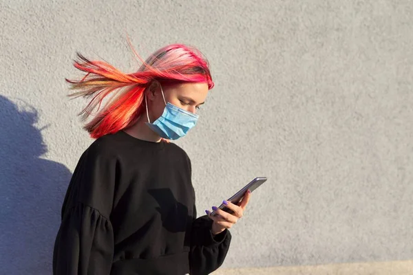 Hipster chica adolescente con máscara de protección médica en la cara, con teléfono inteligente en las manos —  Fotos de Stock