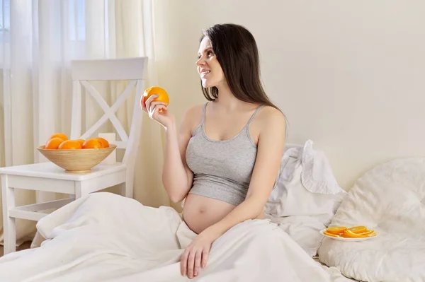 Mladá krásná těhotná žena jíst pomeranče sedí v posteli doma — Stock fotografie